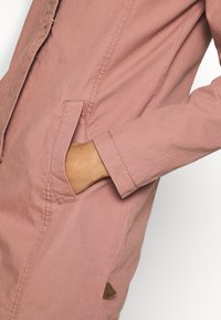 Jasnoróżowa kurtka o gładkiej fakturze, z boczną kieszenią, widocznymi szwami i małą brązową metką z logo przy dolnym brzegu.