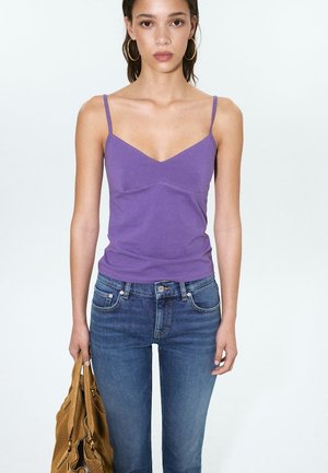 Femme portant un haut violet à fines bretelles et un jean bleu, tenant un sac à main marron dans sa main gauche.