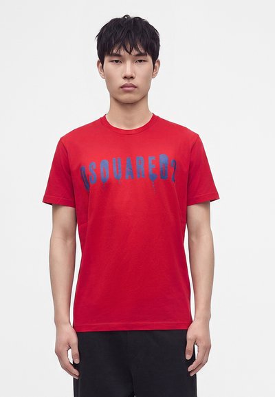 Jeune homme aux cheveux noirs courts portant un T-shirt rouge avec un texte bleu "DSQUARED2" et un pantalon noir, debout sur un fond blanc uni.