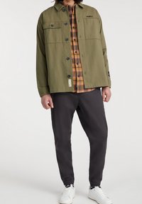 Olivově zelená bunda s knoflíkovou légu a náprsními kapsami, přeložená přes kostkovanou flanelovou košili v žluté a oranžové barvě, doplněná tmavými kalhotami.