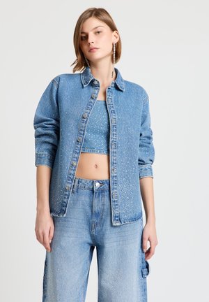 Femme portant une veste en jean bleu clair, un crop top et un jean large avec des détails subtils étincelants, ainsi que de longues boucles d'oreilles pendantes, debout devant un fond blanc.
