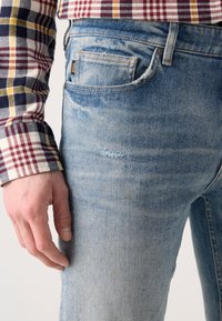 Light blue denim jeans with subtle fading and a distressed patch on the thigh. Hand resting on the fabric, which appears smooth and textured.