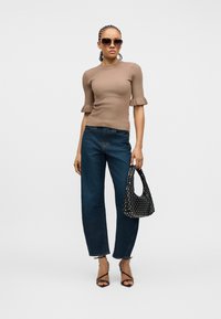 Femme aux cheveux tressés portant des lunettes de soleil, un haut beige à manches froufroutantes, un jean large foncé, un sac noir clouté et des talons noirs à brides.