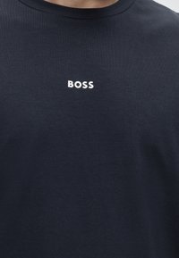 Granatowy t-shirt z okrągłym dekoltem i białym napisem "BOSS" na środku klatki piersiowej, noszony przez osobę.