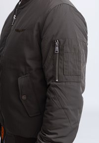 Ciemnozielona kurtka typu bomber o gładkim wykończeniu, z prążkowanymi mankietami, zapinana na zamek kieszeń na rękawie oraz haftowane logo na piersi.