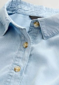 Chemise en denim bleu clair avec un col classique, dotée de cinq boutons de ton contrastant et de détails de couture visibles. Texture douce visible.
