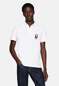 Camisa polo blanca con cuello, logo bordado en el pecho, mangas cortas y tejido texturizado. Se lleva con jeans azul oscuro.