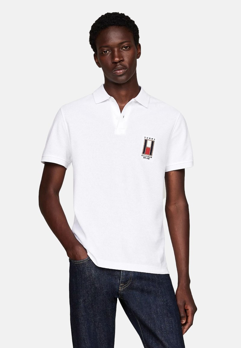 Camisa polo blanca con cuello, logo bordado en el pecho, mangas cortas y tejido texturizado. Se lleva con jeans azul oscuro.