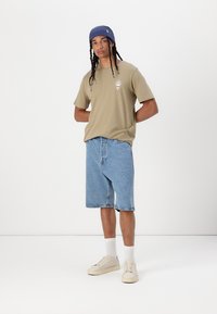 T-shirt kaki à manches courtes avec logo, associé à un short en denim bleu et des baskets blanches. Le modèle porte un bonnet bleu et des chaussettes blanches.