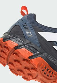 Gros plan d’une chaussure de plein air noire et bleue avec une semelle orange rugueuse, un logo blanc, et un onglet rouge au talon marqué "TERREX."