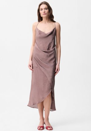 Vrouw draagt een mauve satijnen slipjurk met dunne bandjes en asymmetrische zoom, gecombineerd met rode open tenen hakken, staand tegen een witte achtergrond.