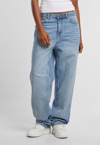 Světle modré džíny z denimu s volným střihem, s knoflíkovým a zipovým zapínáním, pěti kapsami a jemným vyblednutím. Nošeny s bílými teniskami.