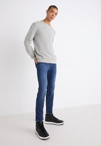 Maglione a maglia grigio chiaro, jeans blu e sneakers alte nere con suole bianche. Il modello è in piedi davanti a uno sfondo semplice.