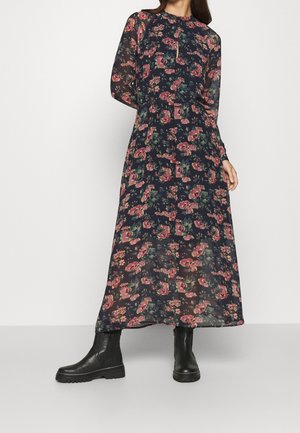 Robe maxi navy avec un motif floral comportant des fleurs roses et vertes. Manches longues, col haut et tissu transparent. Associée à des bottes noires.