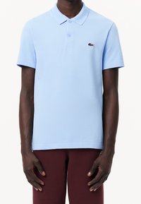Homme portant un polo à manches courtes bleu clair avec un petit logo de crocodile sur la poitrine et un pantalon rouge foncé, debout immobile.