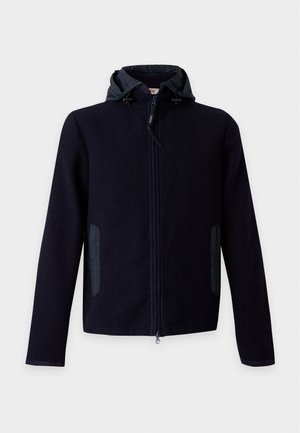 Homme portant une veste noire à capuche, un t-shirt blanc, un pantalon noir et des baskets blanches, debout devant un fond clair uni.