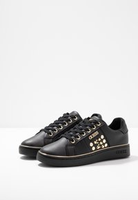 Sneaker nere Guess con borchie dorate e logo sui lati, lacci neri e suole spesse nere con una sottile rifinitura dorata.