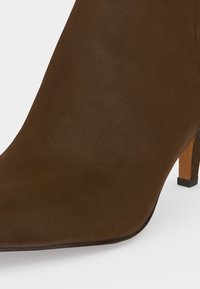 Bottine en daim marron à talon haut avec bout pointu et talon fin sur fond blanc.