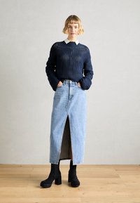 Pull en laine bleu marine à texture épaisse, chemise blanche à col en dessous ; jupe en denim bleu clair avec une fente, bottes à plateforme noires.
