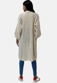 Lange, crèmekleurige gebreide cardigan met een losse pasvorm, verlaagde schouders en een gemêleerd patroon. Draagt over een blauwe jeans en meerkleurige sneakers.