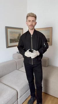 Μαύρο bomber jacket με φερμουάρ μπροστά και τσέπη στο πλάι, συνδυασμένο με μαύρο παντελόνι και λευκά γάντια με σχέδια, κρατημένο μπροστά από ουδέτερο φόντο.