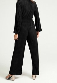 Zwarte, geplooide jumpsuit met lange mouwen, een getailleerde taille en brede benen. Beschikt over een gladde textuur en subtiele verticale lijnen.