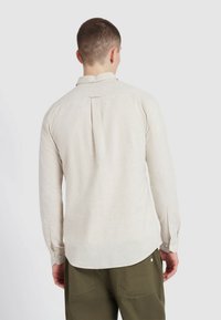 Chemise à manches longues beige clair en tissu texturé, avec un pli dans le dos et des poignets boutonnés, associée à un pantalon vert.
