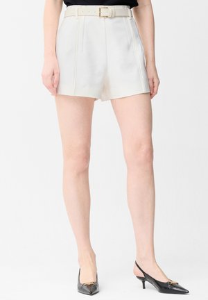 Femme portant un short blanc taille haute sur mesure avec une ceinture, un haut noir et des chaussures slingback noires à petit talon, debout sur fond blanc.