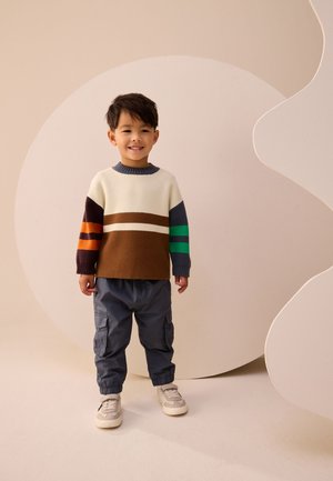 Jeune garçon souriant portant un pull à blocs de couleurs avec des rayures marron, crème, vert et orange, un pantalon cargo gris et des baskets beiges.