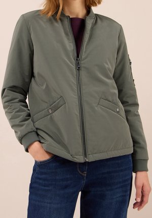 Veste bomber vert olive avec fermeture éclair à l'avant et poches à boutons-pression portée sur un haut violet foncé et un jean bleu.
