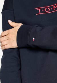 Felpa navy con logo ricamato rosso "T.O.M." sul petto e piccolo accento di bandiera tricolore sul polsino. Tessuto liscio, stile aderente.