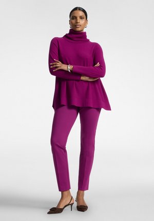 Donna in piedi con le braccia incrociate, indossa un maglione dolcevita magenta, pantaloni aderenti abbinati, un braccialetto d'argento e scarpe marroni con tacco.