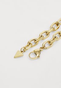 Gouden kettingarmband met onderling verbonden oversized schakels, een driehoekige hanger met gegraveerd logo en een stevige karabijnsluiting.