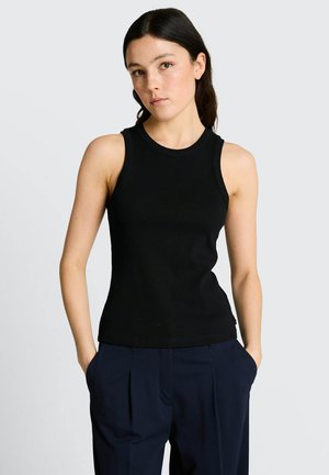 Jonge vrouw met donker haar, gekleed in een mouwloos zwart topje en marineblauwe plooirok, staand met handen in de zakken tegen een effen achtergrond.