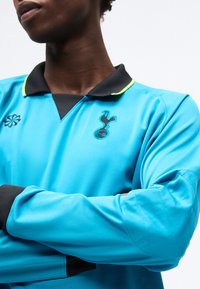 Άτομο που φορά μακρυμάνικη κυανή φανέλα ποδοσφαίρου με μαύρο γιακά και λογότυπο της Tottenham Hotspur, με σταυρωμένα τα χέρια μπροστά σε απλό φόντο.