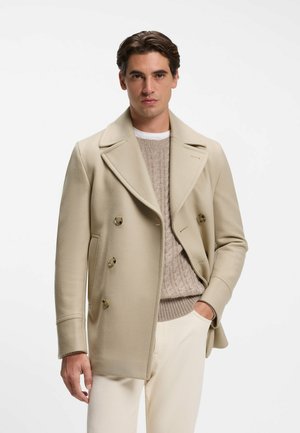 Mann mit dunklen Haaren, der einen beigen zweireihigen Mantel über einem hellbraunen Zopfpullover und offwhite Hosen trägt, Hand in der Tasche, neutraler Gesichtsausdruck.