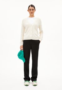 Cremfarbener Cardigan mit Knopfverschluss, schwarze Hose, weißes T-Shirt, hält eine türkisfarbene Kappe und trägt weiße Sneakers mit grünen Akzenten.