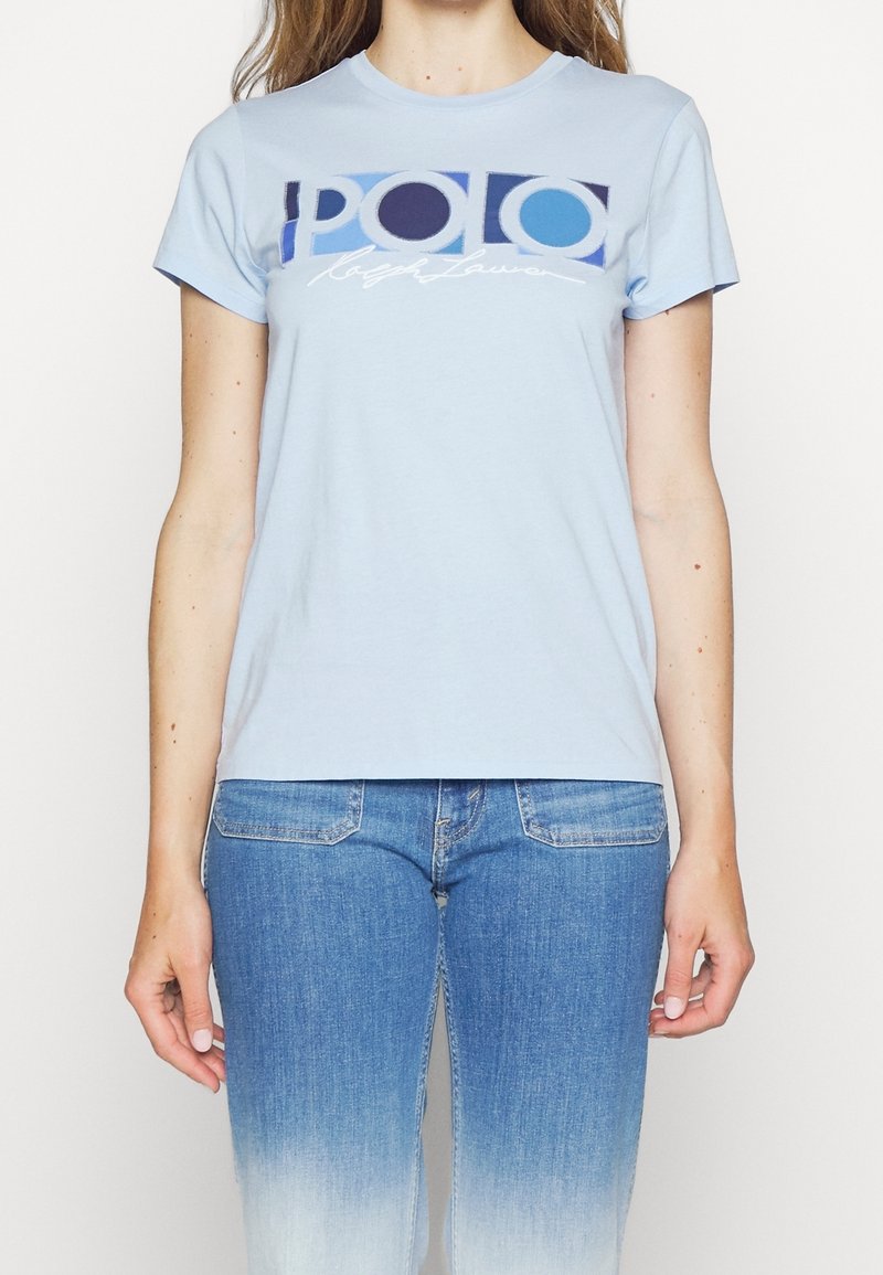 Žena v světle modrém tričku s krátkým rukávem s logem "POLO Ralph Lauren" v modré a bílé barvě, v kombinaci s modrými ombre džíny.