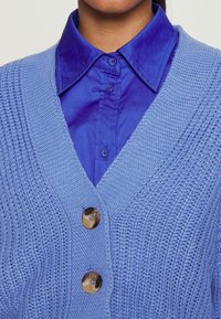 Ljusblå stickad cardigan med V-ringning, prydd med tre stora knappar och texturerad detaljering, bärs över en klarblå skjorta.