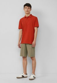 Rode poloshirt met een klassieke kraag, gecombineerd met olivegroene cargoshorts en witte sneakers met bruine accenten. Eenvoudige, casual outfit.