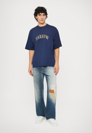 Granatowa, oversize'owa koszulka z napisem "BARROW" w kolorze złotym; zestawiona z wyblakłymi spodniami typu wide-leg z naszywkami i białymi sneakersami.