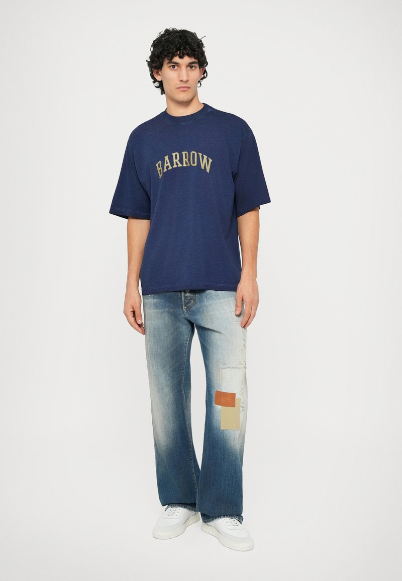 Mørkeblå overdimensjonert t-skjorte med gull "BARROW"-tekst; paret med falmede vide jeans med lappedetaljer og hvite sneakers.