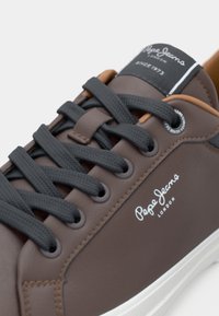Bruna syntetiska sneakers med mörkgrå snören, vit sula, prydda med "Pepe Jeans London"-logotyp och bruna accenter i designen.