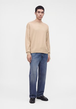 Homme portant une chemise beige à manches longues, un jean bleu et des chaussures noires, debout devant un fond blanc uni.