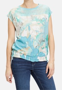 Mujer con una blusa de manga corta de estampado floral turquesa y dobladillo ajustable, combinada con jeans azul oscuro, de pie contra un fondo blanco.