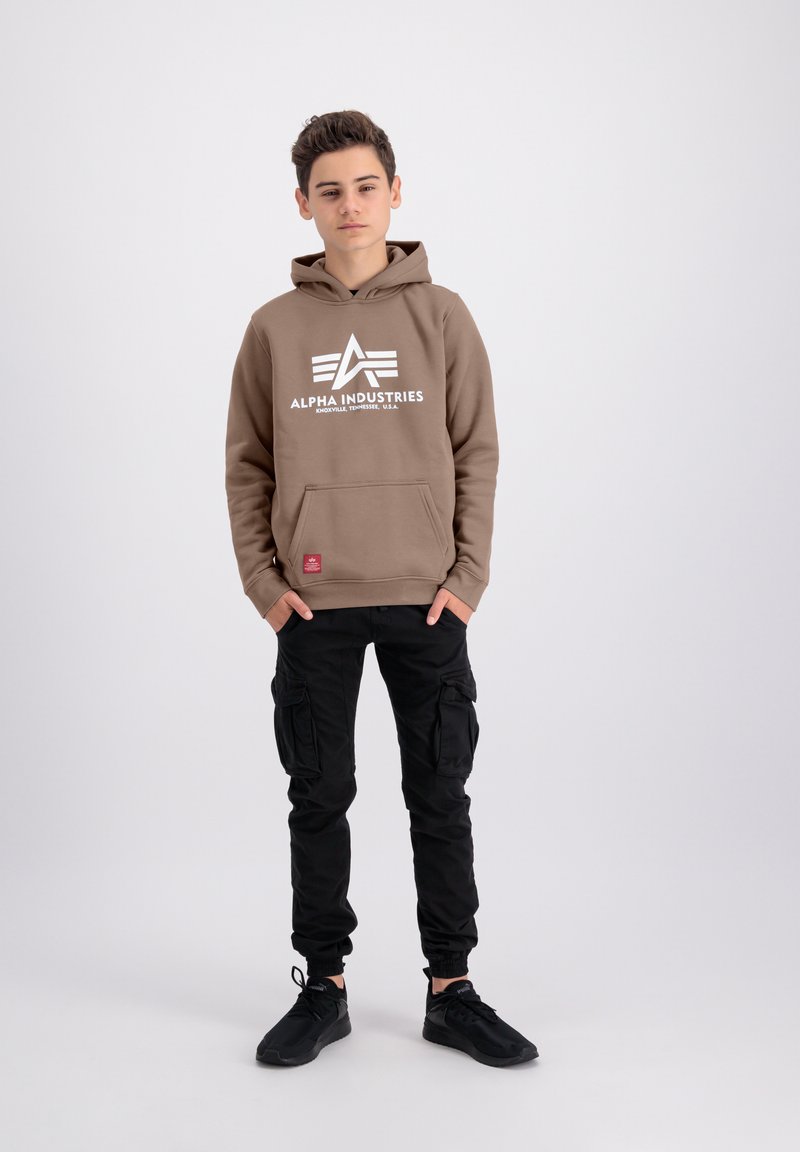 Felpa marrone con logo bianco, tasca frontale e cappuccio con coulisse, abbinata a pantaloni cargo neri e sneakers nere. Un outfit semplice e informale.