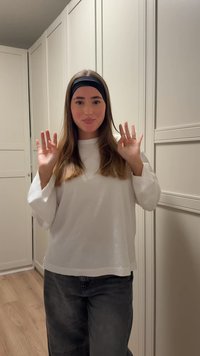 Haut blanc à manches longues avec larges poignets, associé à un jean gris foncé. Sol en bois clair et meubles de cuisine blancs en arrière-plan.