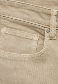 Bež denim tkanina s teksturiranom površinom, koja sadrži prednju džepu sa šivenim rubovima i detaljima od metalnih zakovica.