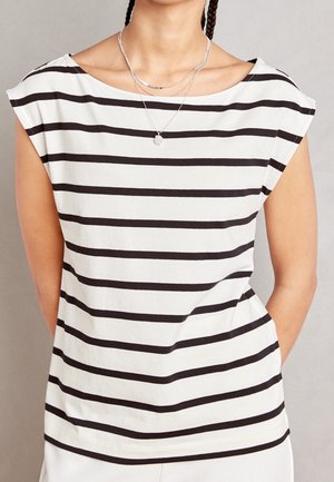 Femme portant un haut blanc sans manches avec des rayures noires horizontales et des colliers argentés superposés, sur un fond neutre.