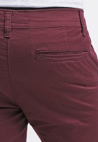 Detailní záběr na osobu v burgundských kalhotách s kapsou na zadní části a viditelným šitím, v kombinaci s bílou košilí.
